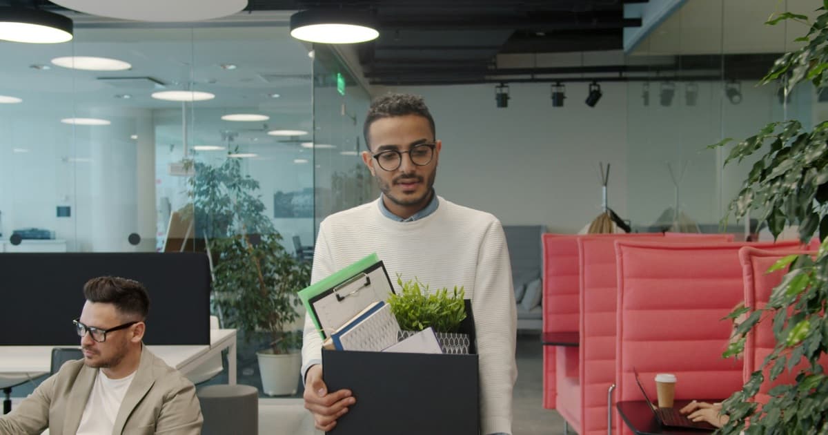 A man carrying a box of his belongings out through an open-plan office on his last day
